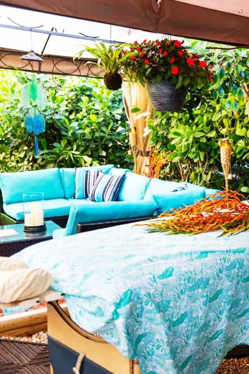 Outdoor Patio With Turquoise Furniture, Patterned Tablecloth, Hanging Plants, And Wind Chimes. Candle And Decorative Items Are Placed On Tables; Lush Green Foliage Surrounds The Patio Area. Outdoor Patio With Turquoise Furniture, Patterned Tablecloth, Hanging Plants, And Wind Chimes. Candle And Decorative Items Are Placed On Tables; Lush Green Foliage Surrounds The Patio Area.