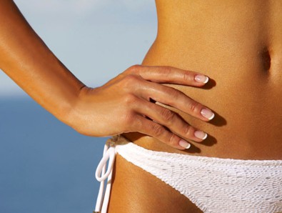 A woman in a white bikini with her hands on her sun-kissed stomach after receiving a professional spray tanning session.